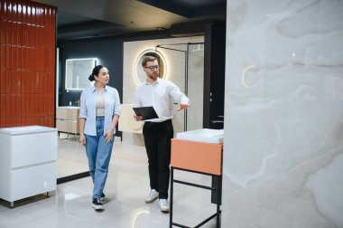 Seramik fayans ve tesisat dükkanını ziyaret eden bir kadın, erkek bir satıcıya ilgi duyuyor. Banyo eşyalar ve tasarım seçenekleri hakkında konuşuyorlar.