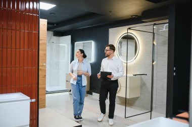 Modern bir tesisat dükkanındaki kadın müşteriye banyo armatürlerini ve fayanslarını gösteren satış temsilcisi.