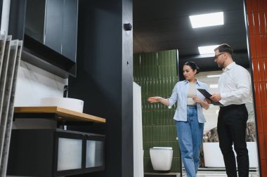 Müşteri, bir su tesisatçısının yeşil fayanslarını gösterip elinde bir pano tutan satıcıyla banyo tasarımını tartışıyor.
