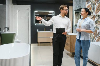 Profesyonel tezgahtar tesisat dükkanında kadınlara banyo demirbaşlarını gösteriyor, satın alma detaylarını tartışıyor.