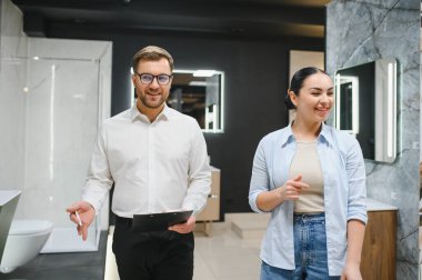 Gülümseyen tezgahtar bir bayan müşteriye farklı banyo armatürlerini gösteriyor, karakteristik özellikleri tartışıyor ve doğru olanı seçmesine yardım ediyor.