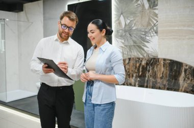 Satıcı, müşteriye tuvaletteki yenileme projesi için seramik fayanslar seçerken bir yandan da panoya not alıyor.