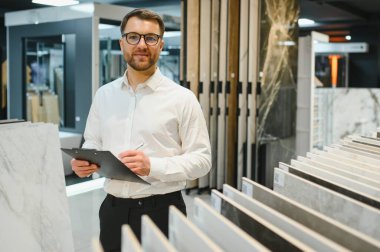 Satış elemanı, seramik fayans ve tesisat mağazasında müşterilere yardım ederken, envanter ve siparişleri yönetirken bir panoya notlar yazıyor.