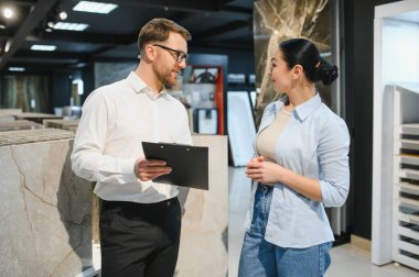 Profesyonel mağaza asistanı elinde bir pano tutuyor. Nalburdaki seramik fayansları seçen kadın müşteriye tavsiye ediyor. Doğru kararı vermesine yardımcı oluyor.