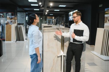 Satış elemanı, parlak, modern bir tesisat mağazasında müşteriye çeşitli seramik fayanslar sunuyor. Tartışıyor ve uzman tavsiyeleri veriyor.