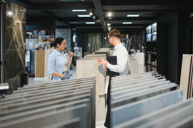 Kadın alıcı, çeşitli fayans ve tesisat vitrinleriyle çevrili modern bir mağazada bir erkek satıcıyla seramik fayans seçeneklerini tartışıyor.