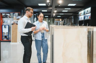 Modern bir sergi salonunda seramik fayansların seçilmesi için kadın müşterilere yardımcı olan profesyonel satıcı, seçenekler ve stiller hakkında tartışmalara girişiyor.