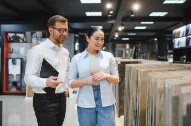 Gülümseyen tezgahtar elinde bir pano var. Tesisat ve fayans dükkanındaki kadın müşteriye seramik fayans örnekleri gösteriyor.