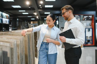 Bir mağazadan seramik fayanslar seçen kadın müşteri, elinde bir pano tutan yardımsever bir satıcı tarafından destekleniyor.