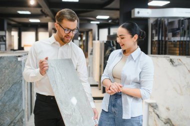 Satıcı, bir tesisat ve fayans dükkanında kadın bir müşteriye seramik fayans örneği gösteriyor.
