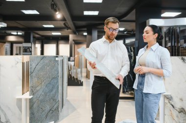 Satıcı, bir kadın müşteriye seramik bir fayans örneği sunuyor. Parlak, modern bir kaplama ve su tesisatı mağazasında. Alışveriş deneyimini geliştiriyor.