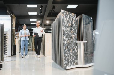 Profesyonel mağaza asistanı pano tutuyor ve hırdavatçıdaki kadın müşteriye farklı seramik fayanslar gösteriyor