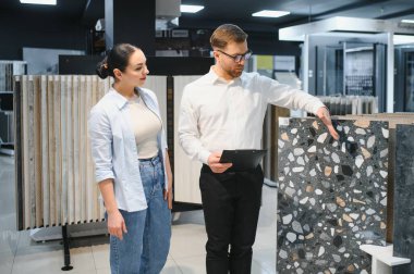 Modern seramik ve su tesisatı mağazasında erkek bir satıcıyla fayans seçeneklerini tartışan kadın müşteri çeşitli fayans tasarımları sergiliyor.