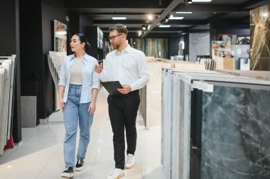 Elinde bir pano olan satıcı, seramik seramik seçenekleri tartışıyor. Hırdavat dükkanının vitrininde yürüyen bir müşteriyle.