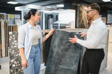 Seramik ve sıhhi tesisat dükkanında seramik fayans sunarken bir kadınla ilişki kuran tezgahtar uzman tavsiyesi ve yardımı sunuyor.