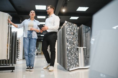 Hırdavatçıda pano tutan satıcıyla konuşurken seramik fayans örneğini gösteren kadın müşteri