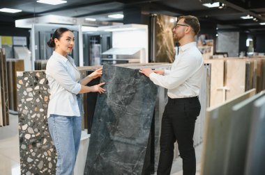 Satış elemanı seramik bir fayans örneğini fayans ve su tesisatı mağazasında bir kadına sunuyor, ev geliştirme seçeneklerini tartışıyor.