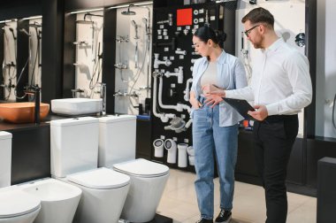 Bir kadın, elinde pano olan bir satıcıyla yeni bir tuvalet seçiyor ve ona farklı modeller hakkında tavsiyelerde bulunuyor.