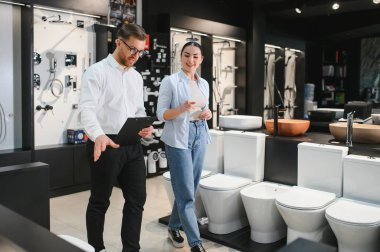 Satıcının elinde bir pano vardı. Tesisatçı dükkanındaki bir kadına farklı tuvaletleri gösteriyordu. Seramik fayansları ve hijyenik eşyaları seçiyordu.