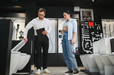 Tesisat ve seramik seramik dükkanında pano tutan kadın müşteriye farklı tuvalet kâseleri gösteren profesyonel bir tezgahtar.