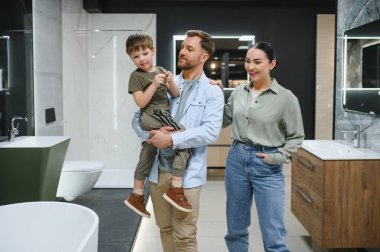 Oğullarını kucağında tutan mutlu ebeveynler bir tesisat dükkanında banyo yenileme için yeni hijyenik malzeme seçiyorlar.