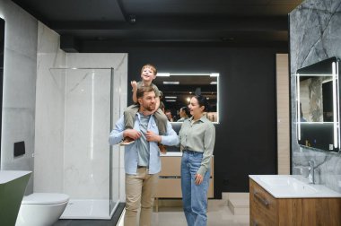 Mutlu bir aile, bir tesisat dükkanında yeni banyo demirbaşlarını seçiyor. Oğullarını omuzlarında taşıyor ve gülümsüyor.