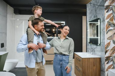 Omuzlarında oğlu olan gülümseyen anne babalar, tesisat dükkanında alışveriş yaparken yeni banyo armatürlerini gösteriyorlar.