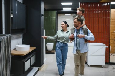Mutlu aile, tesisat dükkanından yeni banyo eşyasına bakıyor. Kadın da lavabo dolabını işaret ediyor.