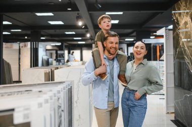 Omuzlarında çocuğu olan gülümseyen bir çift seramik fayans ve su tesisatı dükkanını geziyor, çeşitli fayanslarla çevrili.