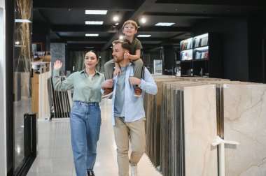 Mutlu aile evlerinin tadilatı için seramik fayansları seçiyor, baba oğlunu omuzlarında taşıyor, anne dükkanda birine el sallıyor.