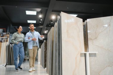 Hırdavatçıda yürüyen genç çift seramik fayanslar seçiyor, tezgahtar onlara çeşitli seçenekler sunuyor.