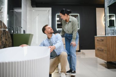 Genç çift gülümseyip tartışırken banyo mobilyaları mağazasında yeni bir küvet seçiyorlar. Birlikte alışverişin tadını çıkarıyorlar.