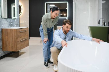 Genç bir çift beyaz bir küveti inceliyor, bir tesisat dükkanında banyo yenilemeyi planlıyorlar.