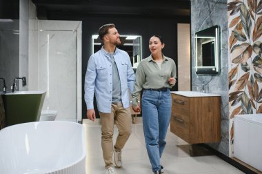 Mutlu çift banyo eşyaları mağazasında el ele yürüyorlar, seramik fayanslar seçiyorlar ve evlerini yenilemek için hijyenik malzeme seçiyorlar.