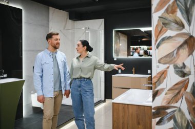 Bir çift el ele tutuşuyor ve bir galeride satış yardımcısıyla konuşurken banyo eşyalar seçiyor.
