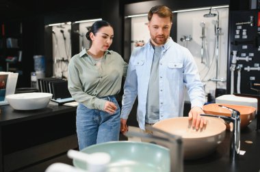 Genç çift seramik lavaboyu bir su tesisatçısında seçiyor, yüzeye dokunuyor ve alışverişi tartışıyorlar.