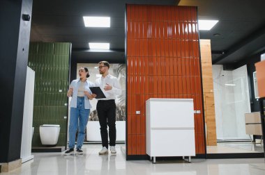 Satıcı elinde seramik ve su tesisatı mağazasındaki kadın müşteriye seramik ve banyo eşyaları sunan bir pano tutuyor.