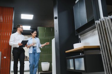 Yeşil seramik fayansları gösteren bir kadın müşteri, bir su tesisatçısında elinde pano tutan bir satıcıyla konuşuyor.