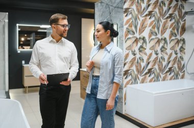 Satıcının elinde bir su tesisatı dükkanındaki kadın müşterilere banyo armatürlerini gösteren bir pano var. Satın alma seçeneklerini tartışıyorlar.