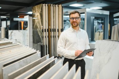 Satıcı, modern bir galeride seramik fayans örneklerini incelerken bir panoya notlar yazıyor, müşteriler için kalite ve çeşitliliği değerlendiriyor.