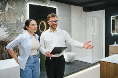 Gülümseyen bir tezgahtar, tesisat mağazasında pano tutan müşterilere banyo mobilyalarını gösteriyor, seramik fayansları ve hijyenik eşyaları seçiyor.