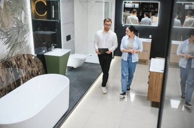 Satıcının elinde modern banyo mobilyası mağazasında yürüyen bayan müşterilere banyo armatürlerini gösteren bir pano var.