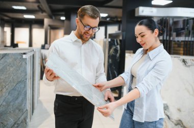 Satış elemanı, bir kadın müşteriye seramik fayans sergileyerek yardımcı oluyor. Parlak, düzenli bir tesisat ve fayans dükkanında. Ev geliştirme seçeneklerini geliştiriyor.