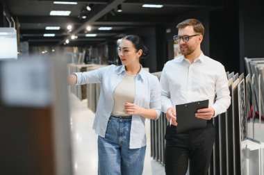 Hırdavatçıda elinde pano tutan satıcıyla alışveriş yaparken seramik fayans örneğine bakan kadın müşteri