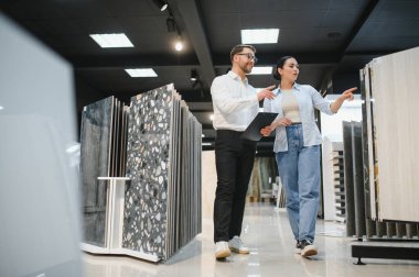 Hırdavatçıdan satış elemanı yardımı alarak seramik fayans örneğini gösteren kadın müşteri, yenileme için tamamlama malzemelerini tartışıyor ve seçiyor