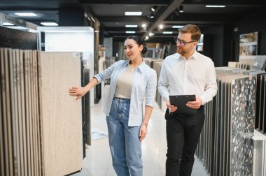Mağazada seramik fayanslar seçen bir kadın ona tavsiye veriyor, elinde bir pano tutuyor ve satın almayı tartışıyor.