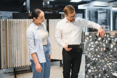 Tezgahtar elinde bir pano tutarken seramik fayans örneğini hareketli bir hırdavatçı ortamında bayan bir müşteriye sunuyor.