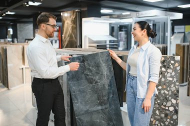 Gülümseyen tezgahtar hırdavatçıda çalışan bir kadına seramik fayans gösteriyor. Yenileme projesi için malzeme seçiyor.