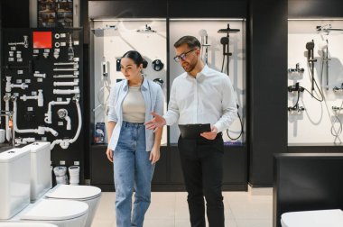 Satış elemanı elinde bir pano ile müşterinin biriyle banyo eşyalar hakkında konuşuyor. Parlak, düzenli bir tesisat galerisinde.