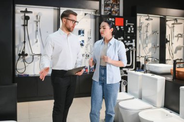 Satış elemanı elinde bir banyo mobilyası dükkanındaki kadın müşteriye farklı klozetler gösteren bir pano tutuyor.